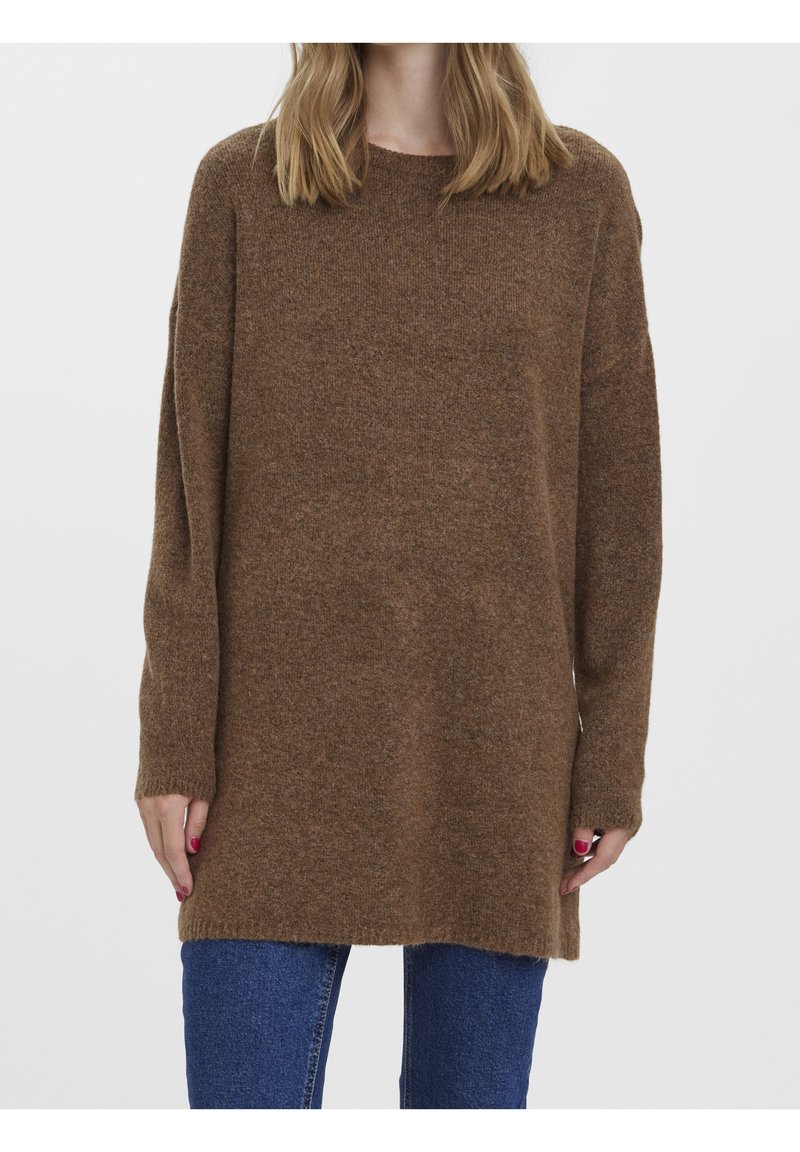 Femme portant un long pull en maille marron avec un col rond et un jean bleu, debout devant un fond clair uni.