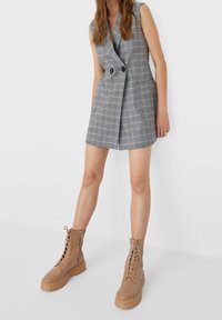 Vestido sin mangas de cuadros grises con diseño cruzado y doble botonadura, con dos bolsillos delanteros, acompañado de botas militares marrones con cordones.