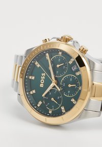 Montre en or et argent avec un cadran vert foncé, présentant trois sous-cadrans, des accents en diamants et un affichage de date. Bracelet en acier inoxydable.