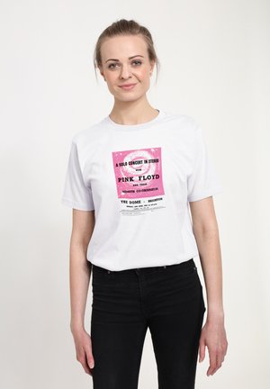Ung kvinde iført en hvid T-shirt med en pink og sort Pink Floyd koncertplakat grafik, kombineret med sorte bukser.