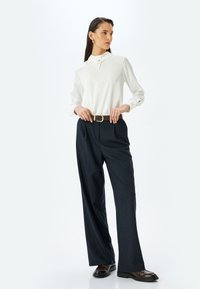 Weiße Bluse mit langen Ärmeln, Kragen und Knopfdetails, kombiniert mit dunkelblauen, gestreiften, weiten Hosen und einem braunen Gürtel. Schwarze Schuhe.