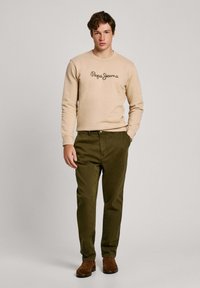Sudadera beige con logo de "Pepe Jeans" en marrón oscuro, combinada con pantalones chinos verde oliva y botines de ante marrón. Conjunto sencillo y casual.