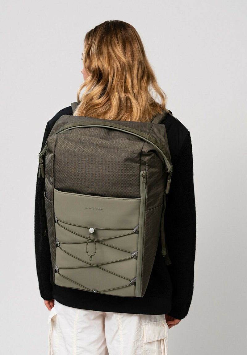Sac à dos vert olive en tissu texturé, avec des compartiments zippés et un détail en cordon élastique sur le devant. Bretelles réglables pour plus de confort.