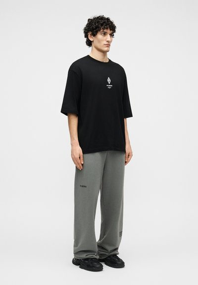 Νέος άνδρας με σγουρά μαλλιά στέκεται φορώντας μια μαύρη φαρδιά μπλούζα με λευκό λογότυπο και φαρδιά γκρι φόρμα με μαύρα αθλητικά παπούτσια.