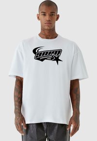 Hvit oversized t-skjorte med korte ermer, med et stort svart grafisk logo og stjerne-design i midten. Glatt bomullstekstur.