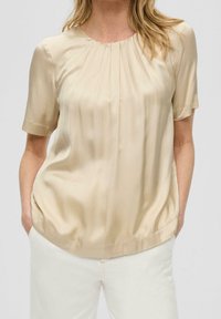 Blouse en satin beige à manches courtes, col rond, plis subtils à l'empiècement et coupe décontractée, associée à un pantalon de couleur claire.