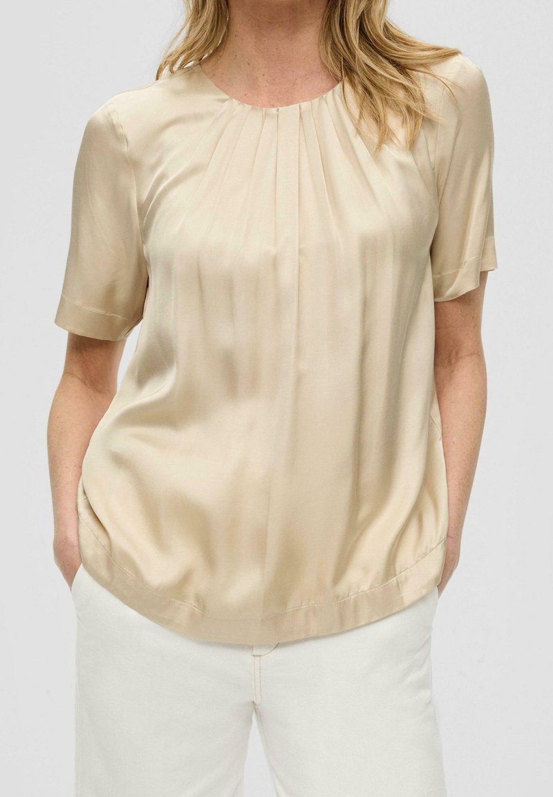 Blouse en satin beige à manches courtes, col rond, plis subtils à l'empiècement et coupe décontractée, associée à un pantalon de couleur claire.