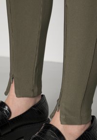 Zbliżenie na prążkowane oliwkowe spodnie z zamkami na kostkach, połączone z czarnymi, teksturowanymi mokasynami na jasnym tle.