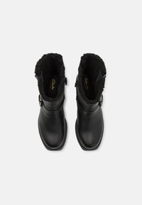 Botas de tobillo de cuero negro con punta redondeada, que cuentan con un forro suave de pelaje negro y una hebilla decorativa en el lado.