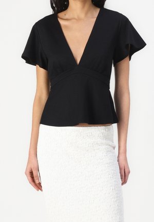 Woman wearing a black, short-sleeve V-neck blouse with gathered waist and a white textured skirt against a plain background.