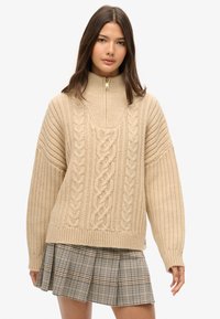 Beige gebreide trui met hoge kraag en rits, voorzien van kabelbrei- en ribbelstructuren. Gecombineerd met een geruite plissérok.