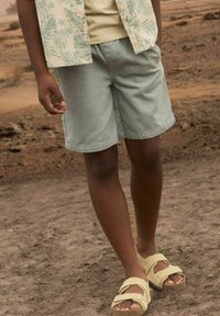 Shorts vert clair à coupe décontractée, associés à une chemise beige avec un motif de feuilles, et des sandales jaunes à deux lanières.
