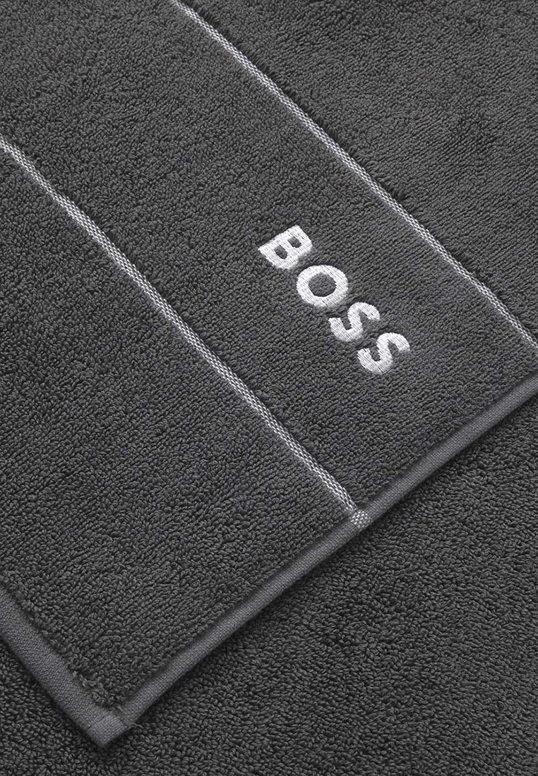 Szary ręcznik o teksturowanej, puszystej powierzchni. Posiada białe haftowane logo "BOSS" oraz kontrastujące obszycia. Prostokątny kształt.