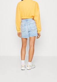 Pantaloni corti in denim azzurro chiaro con orli sfrangiati, due tasche posteriori, abbinati a un pullover giallo corto e sneakers bianche. Sfondo semplice.