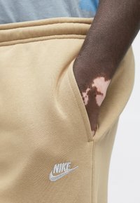 Σκούροχνο χέρι με λεύκη που ξεκουράζεται στην τσέπη μπεζ αθλητικών παντελονιών Nike με λευκό κεντημένο λογότυπο.