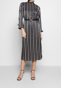 Vestido negro con puntos rosas verticales, de mangas largas, con puños volados, cuello alto, cintura ceñida con un cinturón negro, de longitud midi, con una textura de satén suave.