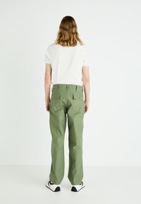 Green cotton pants with a straight leg, featuring side pockets and two back pockets. Paired with a plain white t-shirt and black sneakers.