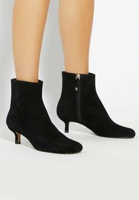 Black suede ankle boots with a pointed toe, short kitten heel, and a side zipper featuring gold hardware accents.