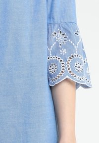 Light blue denim top featuring intricate white embroidered lace at the cuffs, with a scalloped edge and floral patterns.