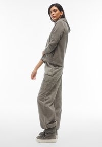 Felpa grigia chiara con cappuccio e pantaloni cargo abbinati, realizzati in tessuto morbido e testurizzato. La felpa ha un cordino mentre i pantaloni sono dotati di tasche.