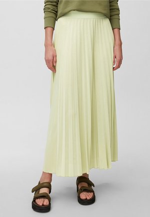 Persoon die een lichtgele geplooide lange rok, een olijfgroene trui en olijfgroene sandalen met dikke zwarte zolen draagt.
