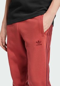 Pantalones deportivos rojos hechos de tela suave, con tres franjas de color rojo oscuro a lo largo del costado y un logo bordado negro en el muslo.