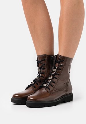 Hnědé kožené kotníkové boty s hladkým povrchem, černým šněrováním vpředu, bočním zipem a texturovanými podrážkami. Disponují stříbrnými kovovými doplňky.