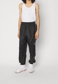 Schwarze Nylon-Trainingshose mit elastischem Bund, Seitenstreifen und kontrastierenden Einsätzen. Kombiniert mit weißen Sneakers mit roten Akzenten.