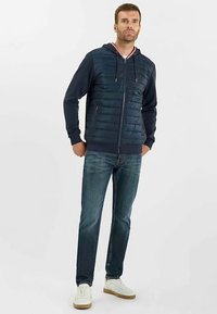 Giacca imbottita blu navy con design uniforme, chiusura con cerniera e cappuccio, abbinata a jeans blu e scarpe da ginnastica bianche, in piedi contro uno sfondo chiaro.