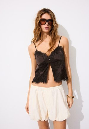 Frau mit welligem Haar trägt übergroße Sonnenbrille, schwarzen Spitzen-Camisole, weiße Shorts mit hoher Taille und goldenen Armreif vor weißem Hintergrund.