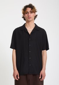 Jeune homme aux cheveux ondulés portant une chemise noire à manches courtes à boutons et un pantalon marron, se tenant devant un fond blanc uni.