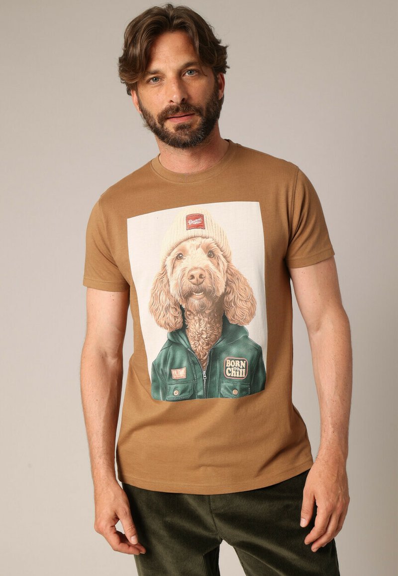 Bruine katoen T-shirt met een vooropdruk van een hond in een groene jas en gebreide muts, met de tekst "BORN CHILL" op de jas.