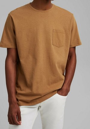 Homme portant un t-shirt marron uni à manches courtes avec une poche poitrine et un pantalon blanc, debout avec une main dans la poche.
