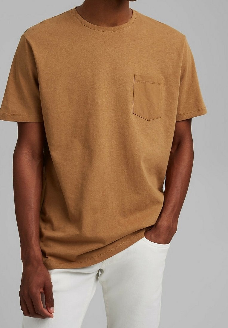 Homme portant un t-shirt marron uni à manches courtes avec une poche poitrine et un pantalon blanc, debout avec une main dans la poche.