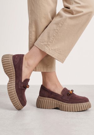 Person iført beige bukser og lilla ruskind platform loafers med chunky rillede gummisåler og træperle detaljer.