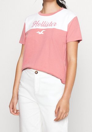 Mujer vestida con una camiseta de Hollister en rosa y blanco, metida dentro de unos pantalones blancos de cintura alta, de pie contra un fondo liso.