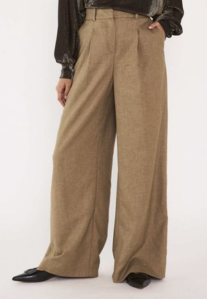 Beige broek met wijde pijpen, hoge taille en ceintuurlussen, gedragen met een glanzend zwart blouse met lange mouwen en zwarte puntige schoenen.