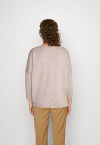 Maglione in maglia oversize beige chiaro con maniche a tre quarti, texture liscia e una semplice cucitura sul retro, abbinato a pantaloni marrone chiaro.
