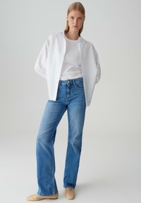 Witte knopenoverhemd, effen wit T-shirt eronder, blauwe rechte jeans, lichtbeige schoenen, tegen een neutrale achtergrond.