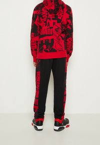 Punainen ja musta kuviollinen huppari, jossa on pitkät hihat ja huppu, yhdistettynä mustiin collegehousuihin. Materiaalit näyttävät pehmeiltä ja teksturoiduilta.