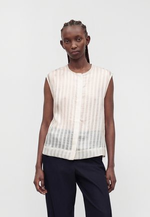 Femme aux cheveux tressés portant un chemisier blanc transparent sans manches à rayures et un pantalon noir, debout devant un fond clair uni.