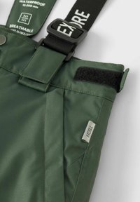 Žalios vandeniui atsparios kelnės su reguliuojamais juodais petnešomis. Ypatybės: lipduko skląstis, audinio etiketas ir lygus, kvėpuojantis paviršius.