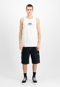 Λευκό αμάνικο μπλουζάκι με το λογότυπο "Alpha Industries" και μαύρες cargo βερμούδες με αντίθετες λευκές ραφές και πολλές τσέπες, συνδυασμένες με μαύρα αθλητικά παπούτσια.