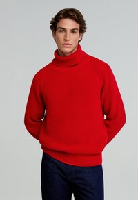 Suéter de cuello alto de canalé rojo con un ajuste holgado, que presenta un cuello alto y mangas raglán, combinado con jeans azul oscuro.