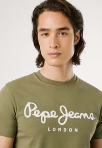 Camiseta de manga corta verde oliva de algodón, que presenta un gran logo blanco "Pepe Jeans LONDON" en el pecho en una fuente cursiva.