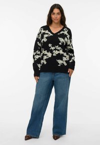 Zwarte trui met wit bloemenpatroon, V-hals ontwerp, oversized pasvorm, gecombineerd met wijde blauwe jeans en puntige bruine schoenen.