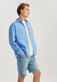 Chemise rayée bleu clair avec une coupe décontractée, portée par-dessus un T-shirt blanc, associée à un short en denim déchiré bleu clair.
