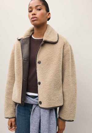 Le mannequin porte une veste beige en peau lainée à col foncé par-dessus un haut marron et un jean, et se tient devant un fond de studio uni.