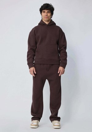 Hoodie marron avec une poche frontale, des poignets et une taille côtelés, associé à un pantalon de survêtement assorti. Le matériau semble doux et comme en polaire.