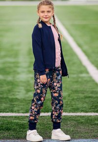 Młoda dziewczyna z warkoczami, ubrana w kwiatowe spodnie, granatową kurtkę i białe trampki, stoi na boisku sportowym obok białej linii.